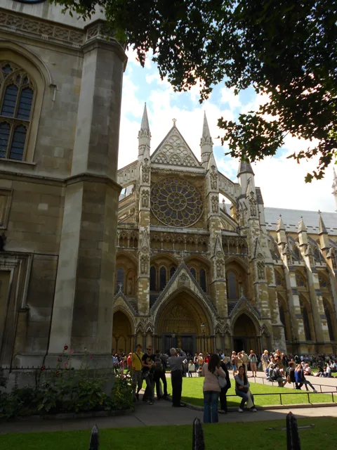 People walking beside a stone abbey under leafy trees.