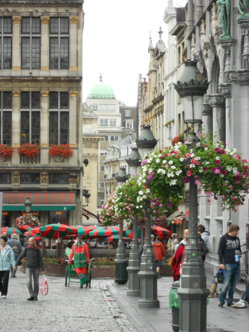 Pedestrians on a cobblestone street beside striped café umbrellas and hanging flowers.